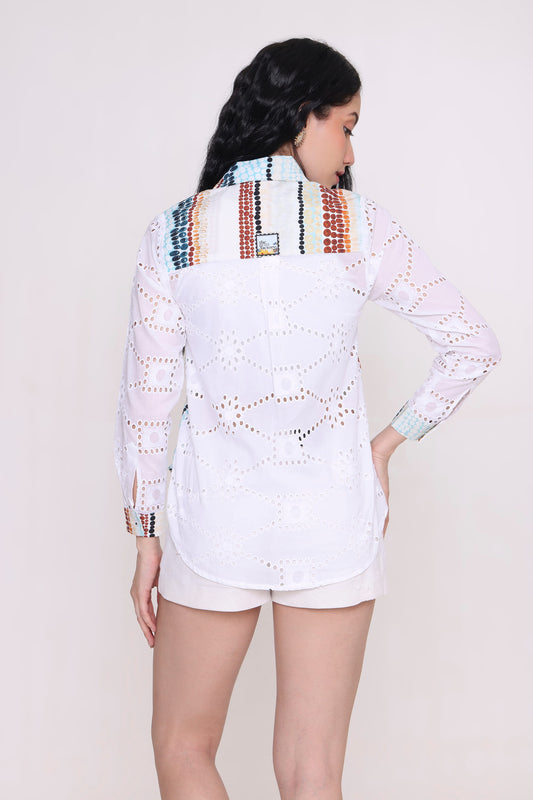A woman shown from the back wearing a white long-sleeve eyelet shirt with colourful geometric embroidery across the shoulders and a white shorts, against a plain light beige studio background.