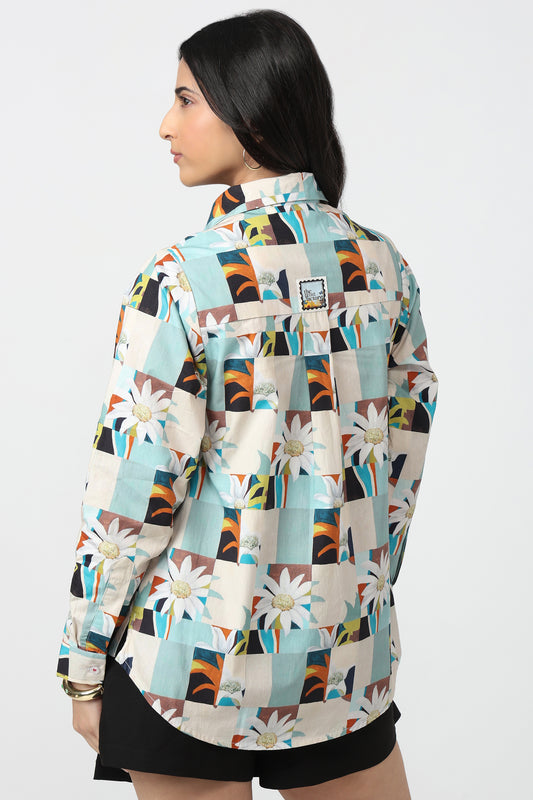 A woman shown from the back wearing a long-sleeve button-up shirt with a colorful geometric patchwork print featuring daisies, abstract shapes, and blocks of teal, orange, black, and cream.