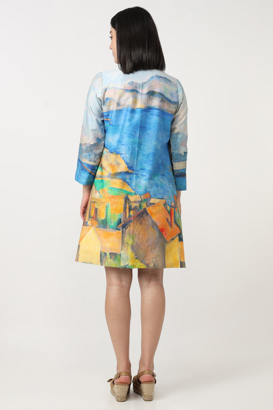 A woman viewed from the back wearing a short-sleeve A-line dress with a colorful landscape print in shades of blue, yellow, green, and orange, set against a plain light gray studio background.