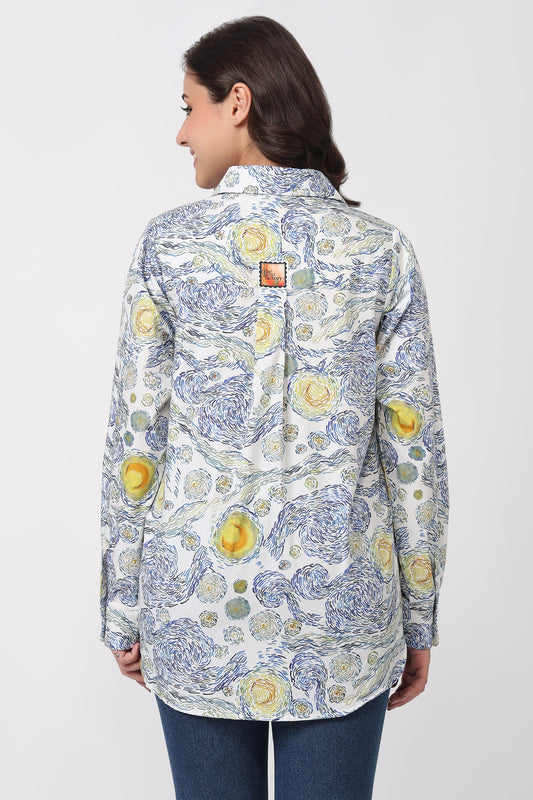 A woman shown from the back wearing a long-sleeve button-up shirt with a blue and yellow floral toile-style print on a white background, paired with blue jeans against a light gray studio background.