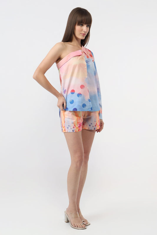 A woman modeling a pastel one-shoulder wrap top and matching shorts set with a pink, blue, and orange watercolor-style print and polka dots, set against a plain white studio background.