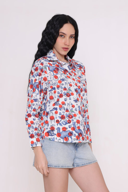 A woman modeling a white long-sleeve button-up shirt with a red and blue cherry print, paired with light-wash denim shorts, against a plain light background.