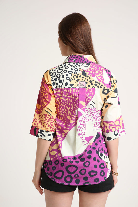A woman shown from the back wearing a short-sleeve button-up shirt with a bold multicolor animal print pattern in pink, purple, orange, black, and white, paired with black shorts against a light gray studio background.