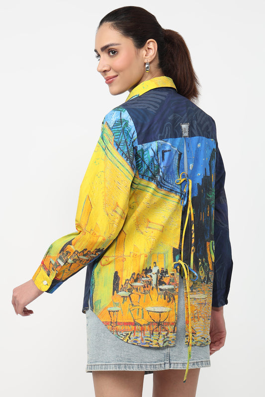 A woman modeling a long-sleeve button-up shirt with a Van Gogh-inspired cafe print, featuring yellow, blue, and navy panels, paired with a denim skirt against a white studio background.