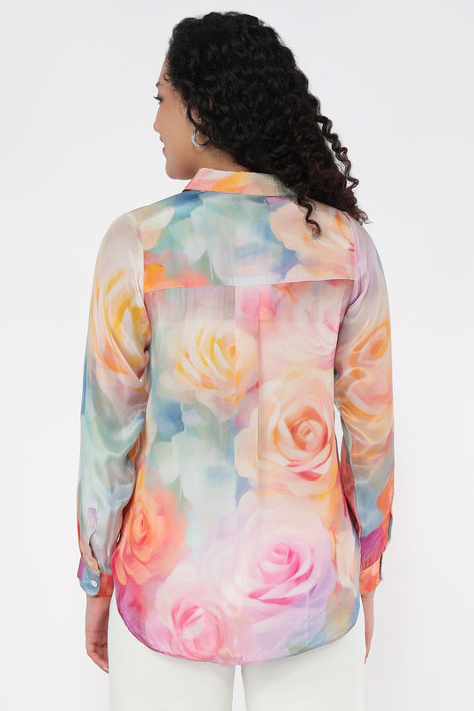 A woman viewed from the back wearing a sheer long-sleeve button-up shirt with a pastel watercolour floral print featuring pink, peach, yellow, and blue tones, paired with white pants against a light grey studio background.