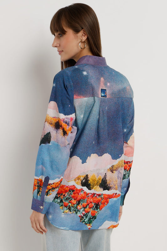 A woman shown from the back wearing a long-sleeve button-up shirt with a colorful landscape print featuring blue skies, mountains, and red flowers, paired with light gray jeans against a plain light gray background.
