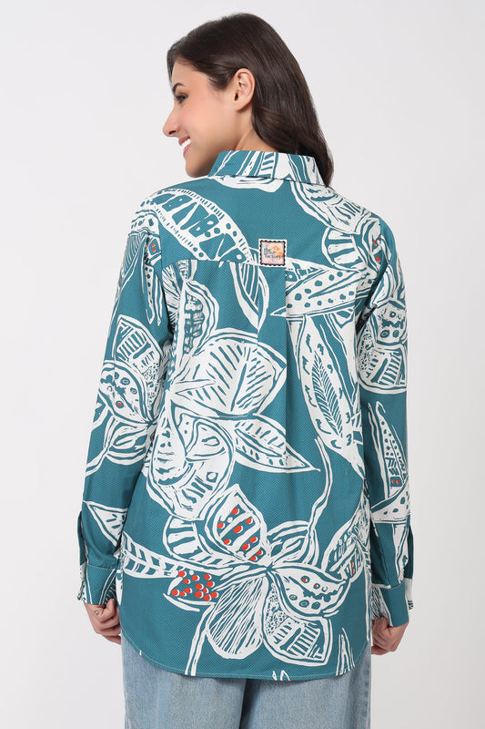 A woman shown from the back wearing a teal button-up shirt with a large white tropical leaf pattern and light blue jeans, against a plain light gray studio background.