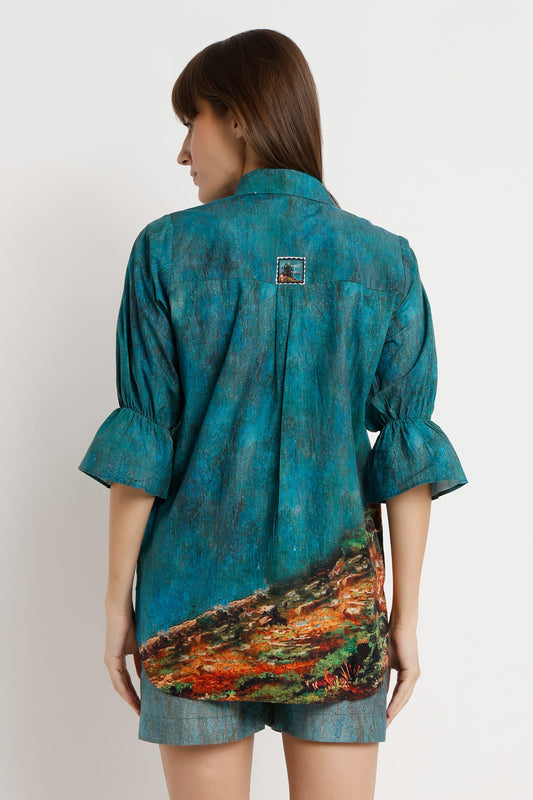 A woman shown from the back wearing a teal blue short-sleeve shirt with a colorful abstract landscape print on the lower back and denim shorts, against a plain light background.