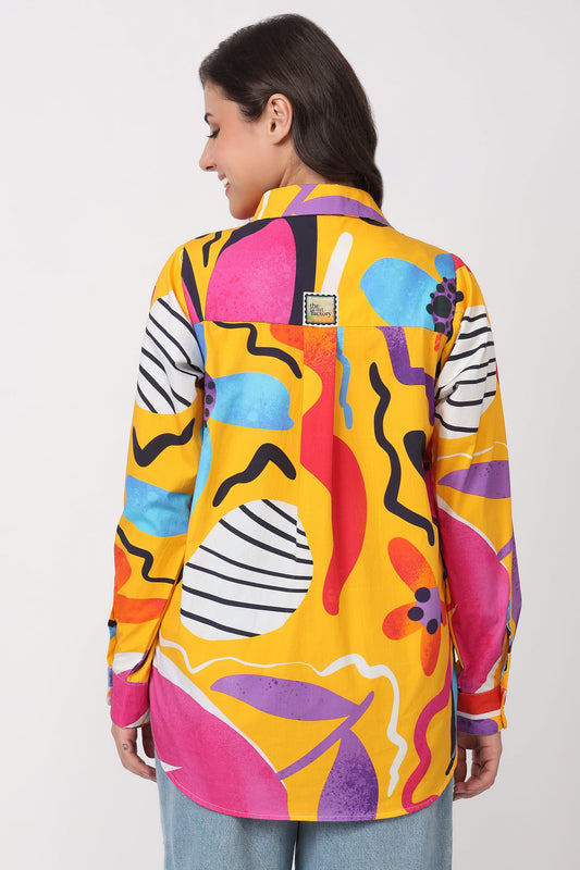 A woman shown from the back wearing a long-sleeve button-up shirt with a bold, multicolored abstract print on a yellow base, paired with light blue jeans against a plain light gray background.