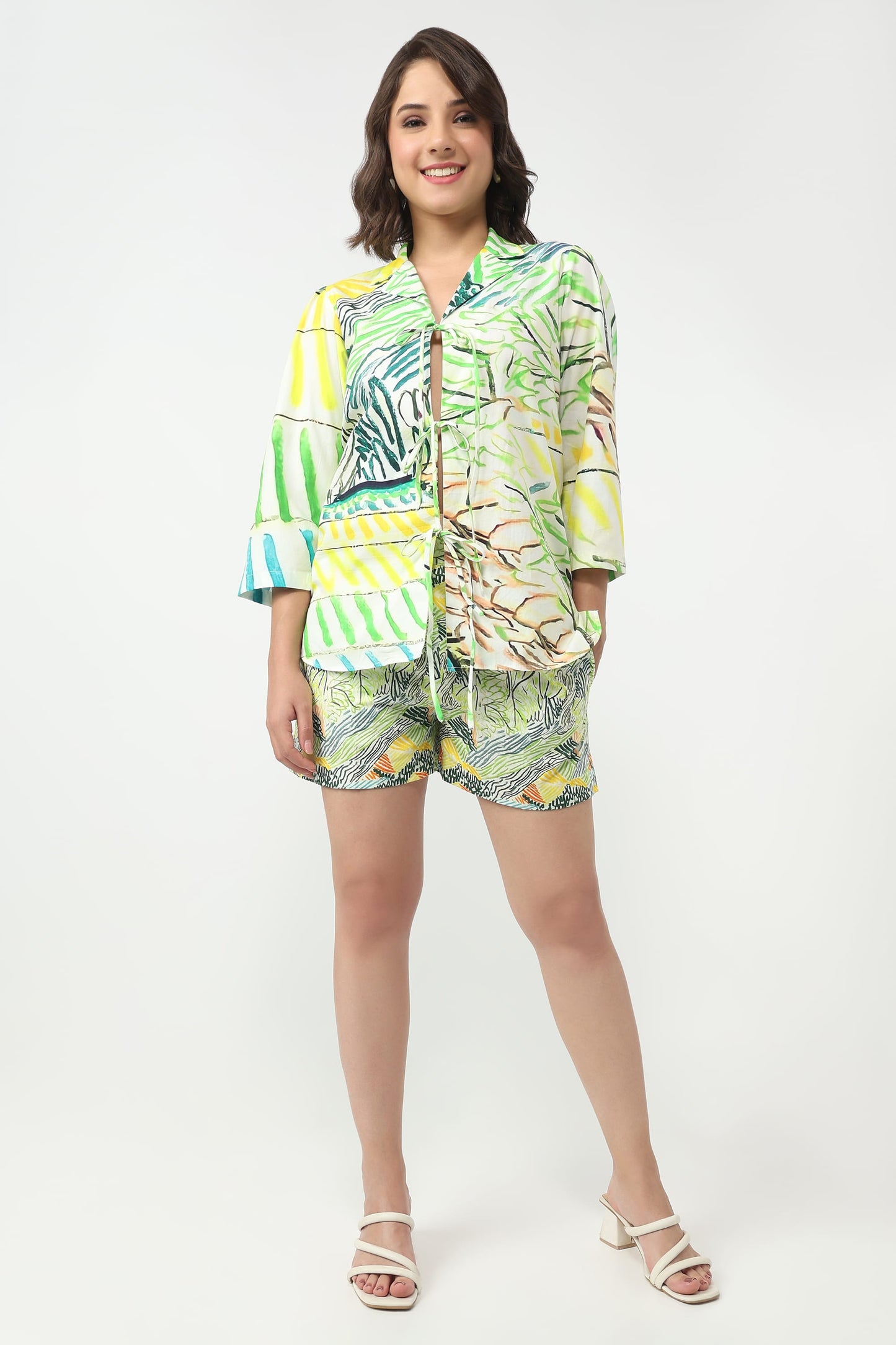 A woman modeling a matching tropical print two-piece set consisting of a short-sleeve button-up shirt and shorts, paired with white heeled sandals, against a plain white studio background.