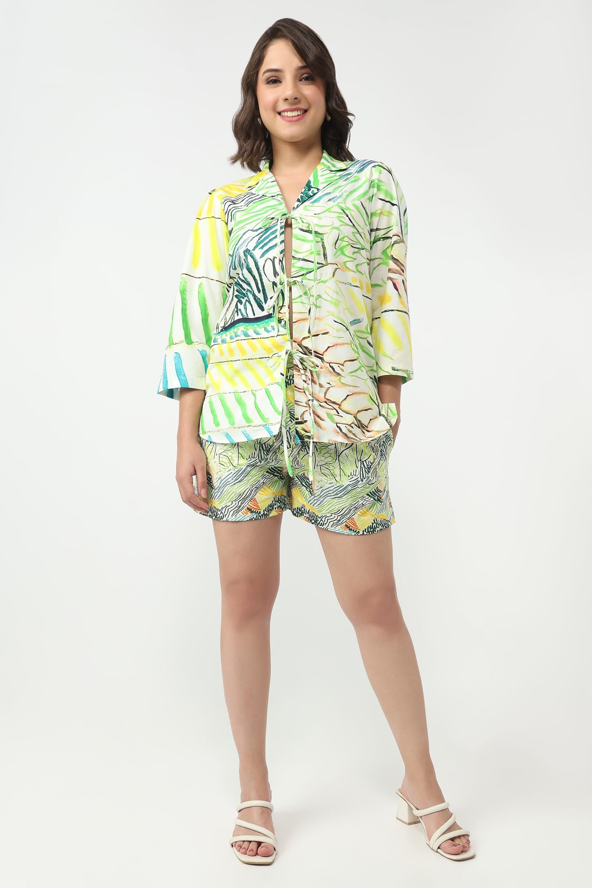 A woman modeling a matching tropical print two-piece set consisting of a short-sleeve button-up shirt and shorts, paired with white heeled sandals, against a plain white studio background.