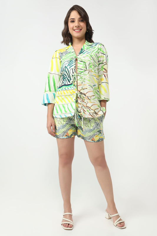 A woman modeling a matching tropical print two-piece set consisting of a short-sleeve button-up shirt and shorts, paired with white heeled sandals, against a plain white studio background.
