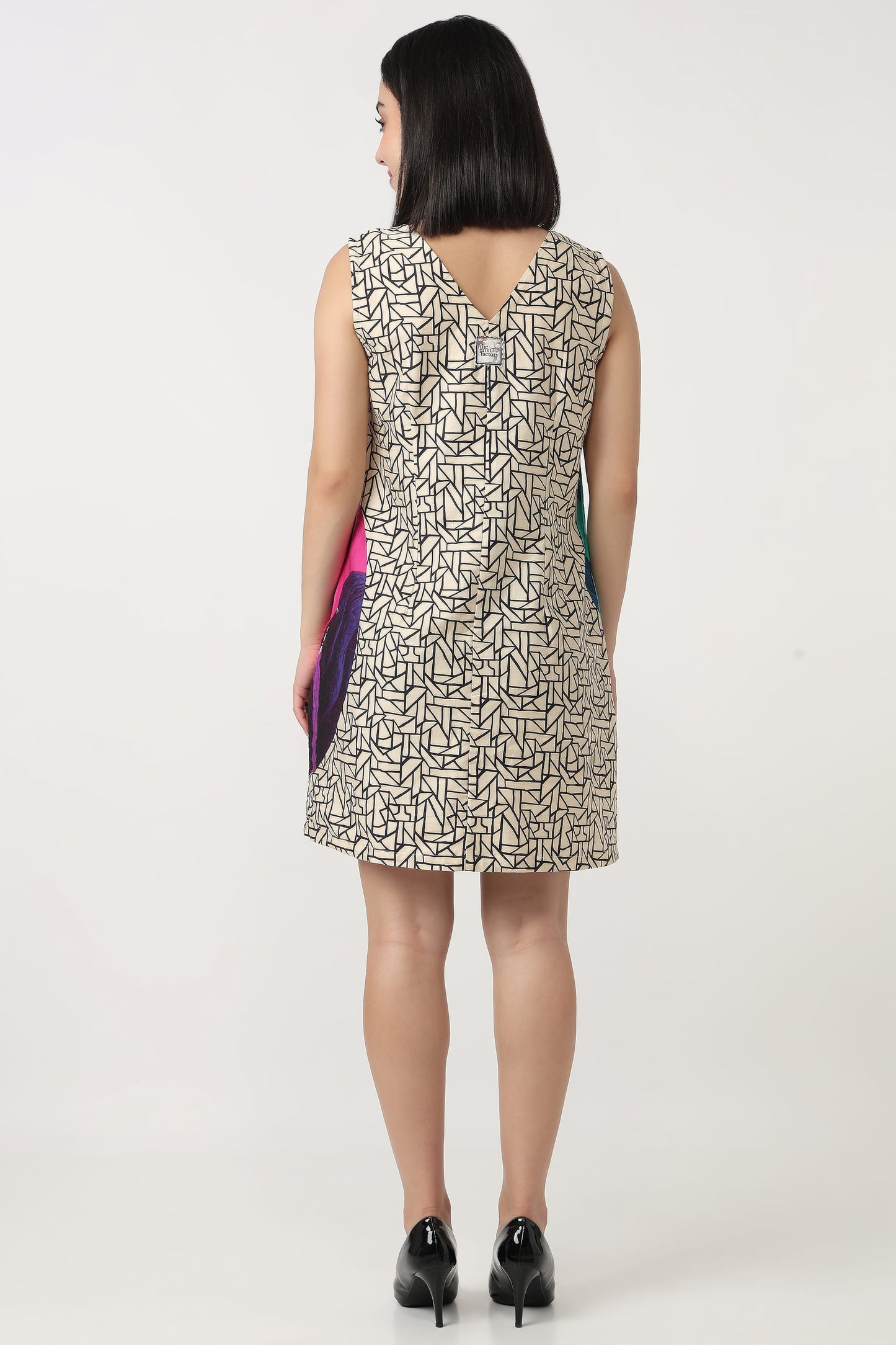 A woman viewed from the back wearing a sleeveless knee-length dress with an abstract black line pattern on a light beige background. She is holding a small colorful clutch and wearing black heels against a plain white studio background.