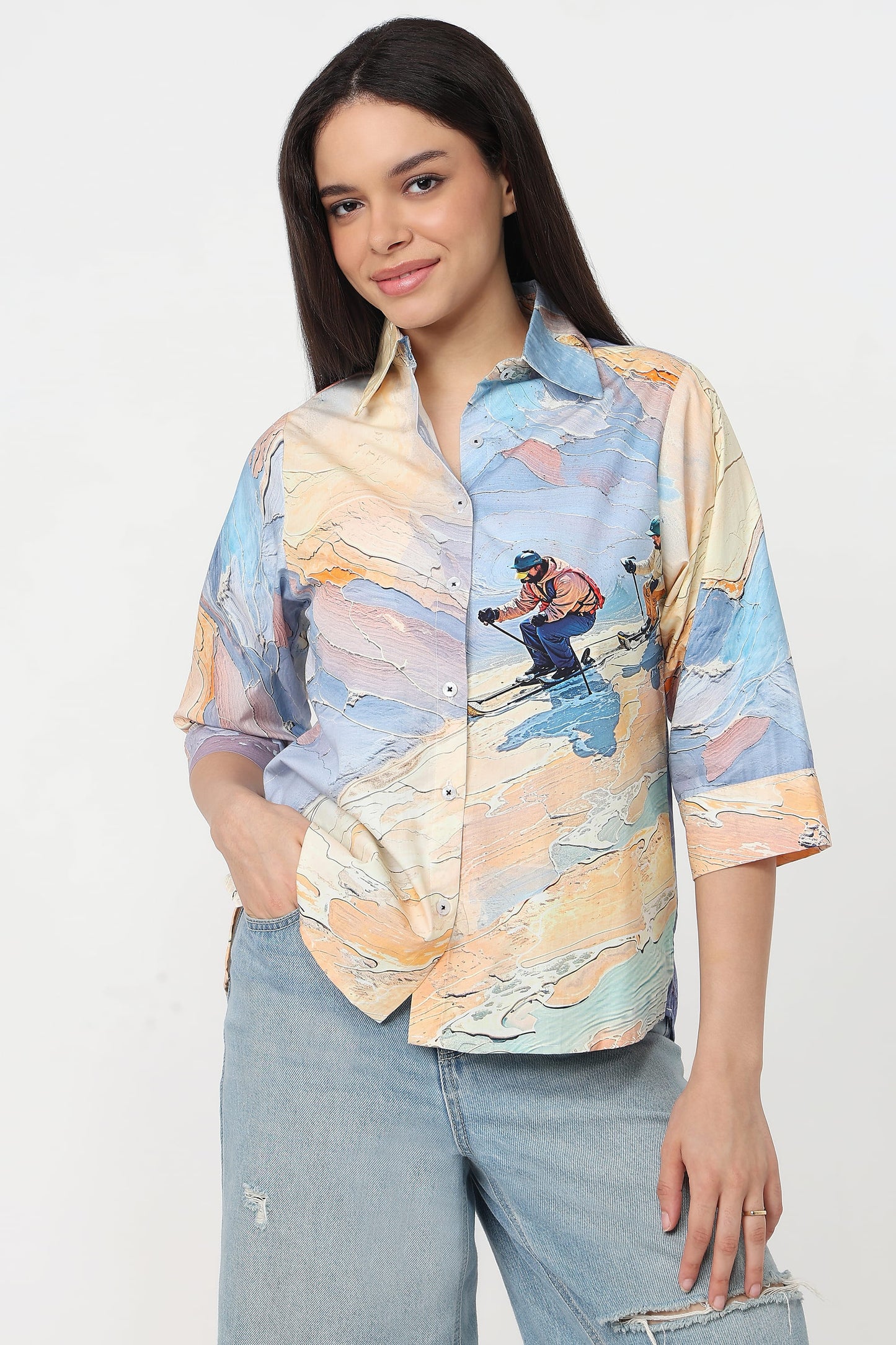 A woman wearing a short-sleeve button-up shirt with a colourful abstract landscape print featuring a skier, in light blue jeans against a plain white studio background.