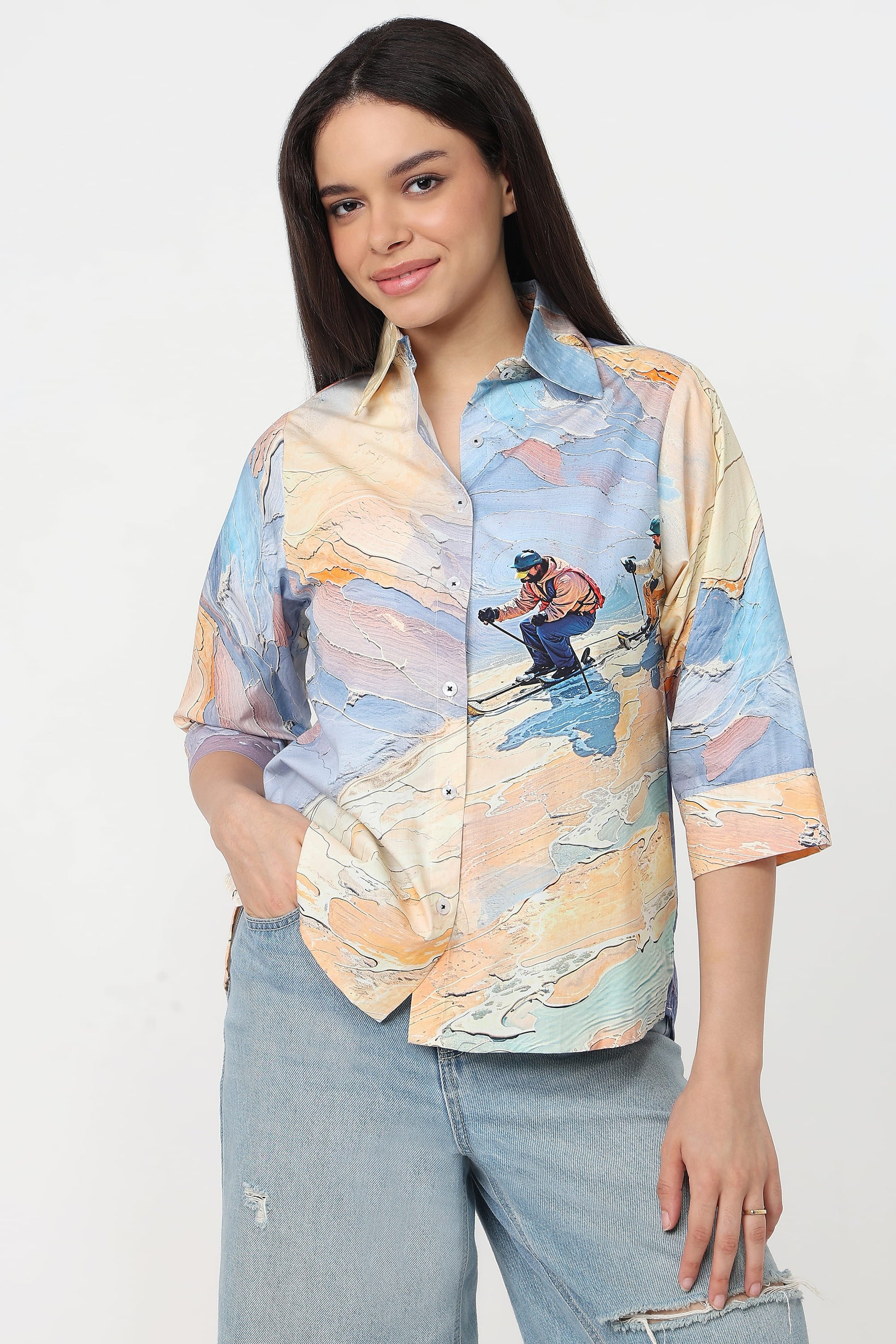 A woman wearing a short-sleeve button-up shirt with a colourful abstract landscape print featuring a skier, in light blue jeans against a plain white studio background.