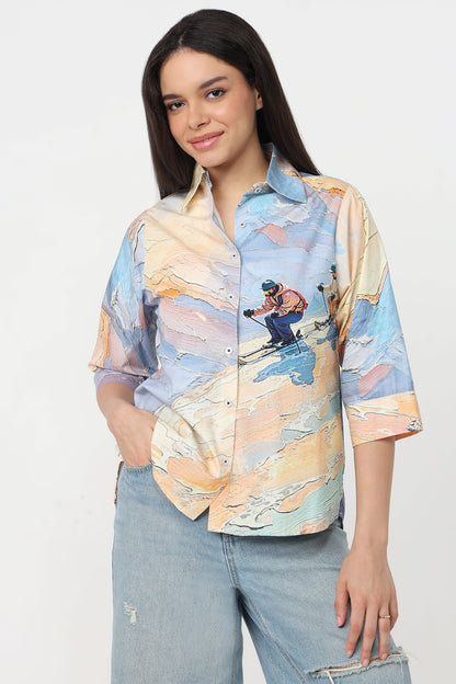 A woman wearing a short-sleeve button-up shirt with a colourful abstract landscape print featuring a skier, in light blue jeans against a plain white studio background.