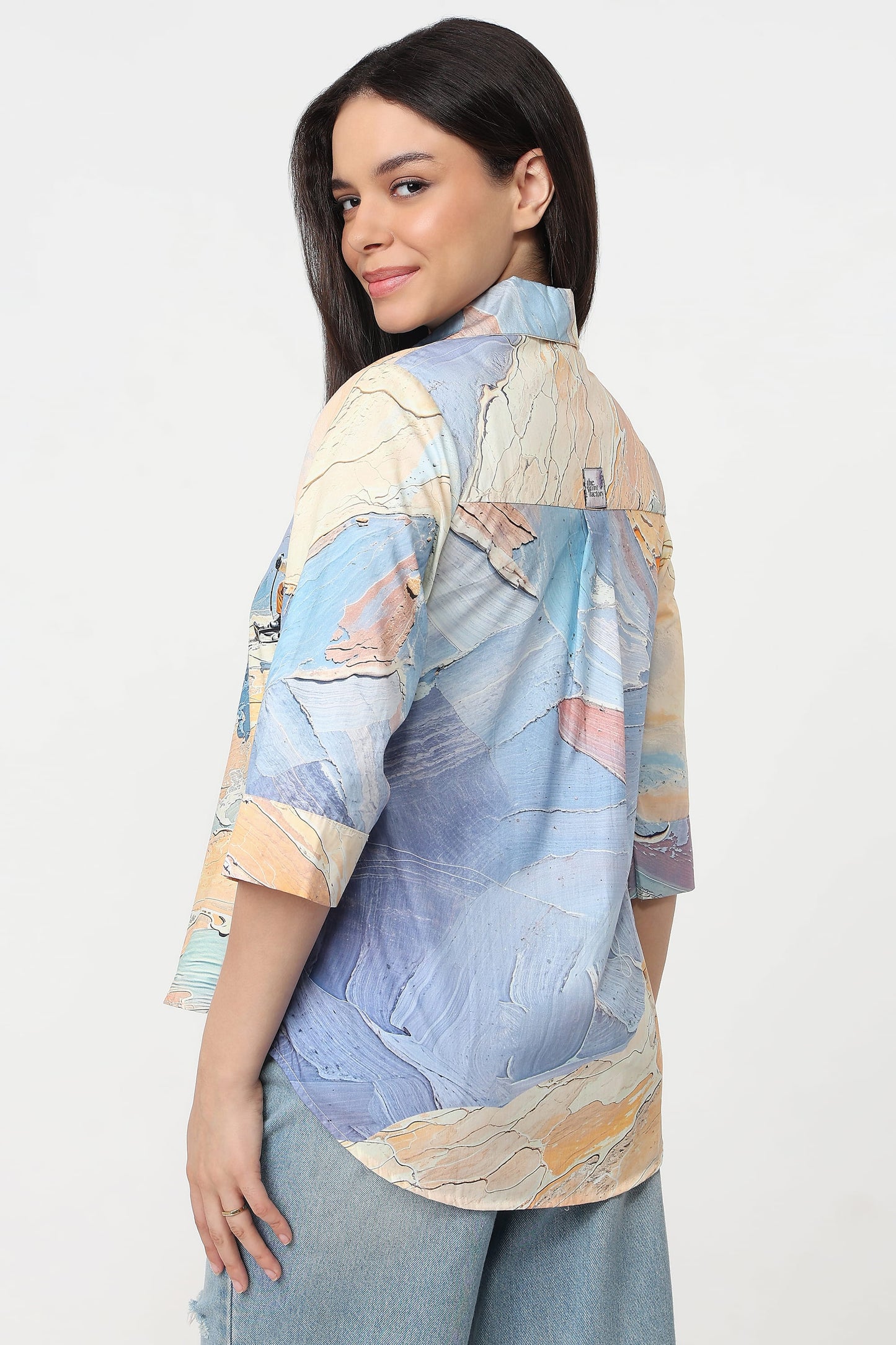 A woman wearing a light blue and beige abstract-patterned button-up shirt with three-quarter sleeves, paired with light-wash jeans, photographed against a plain white studio background.