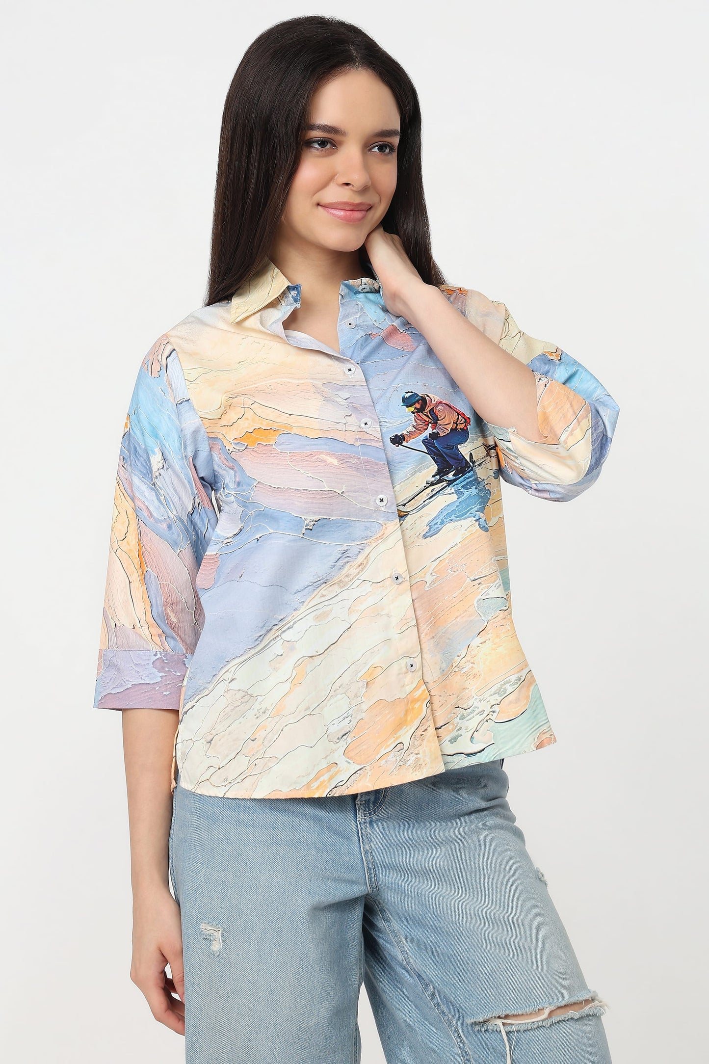 A woman wearing a short-sleeve button-up shirt with a colourful abstract print in shades of blue, beige, and orange, featuring a small skier graphic on the chest, paired with light-wash jeans against a plain white background.