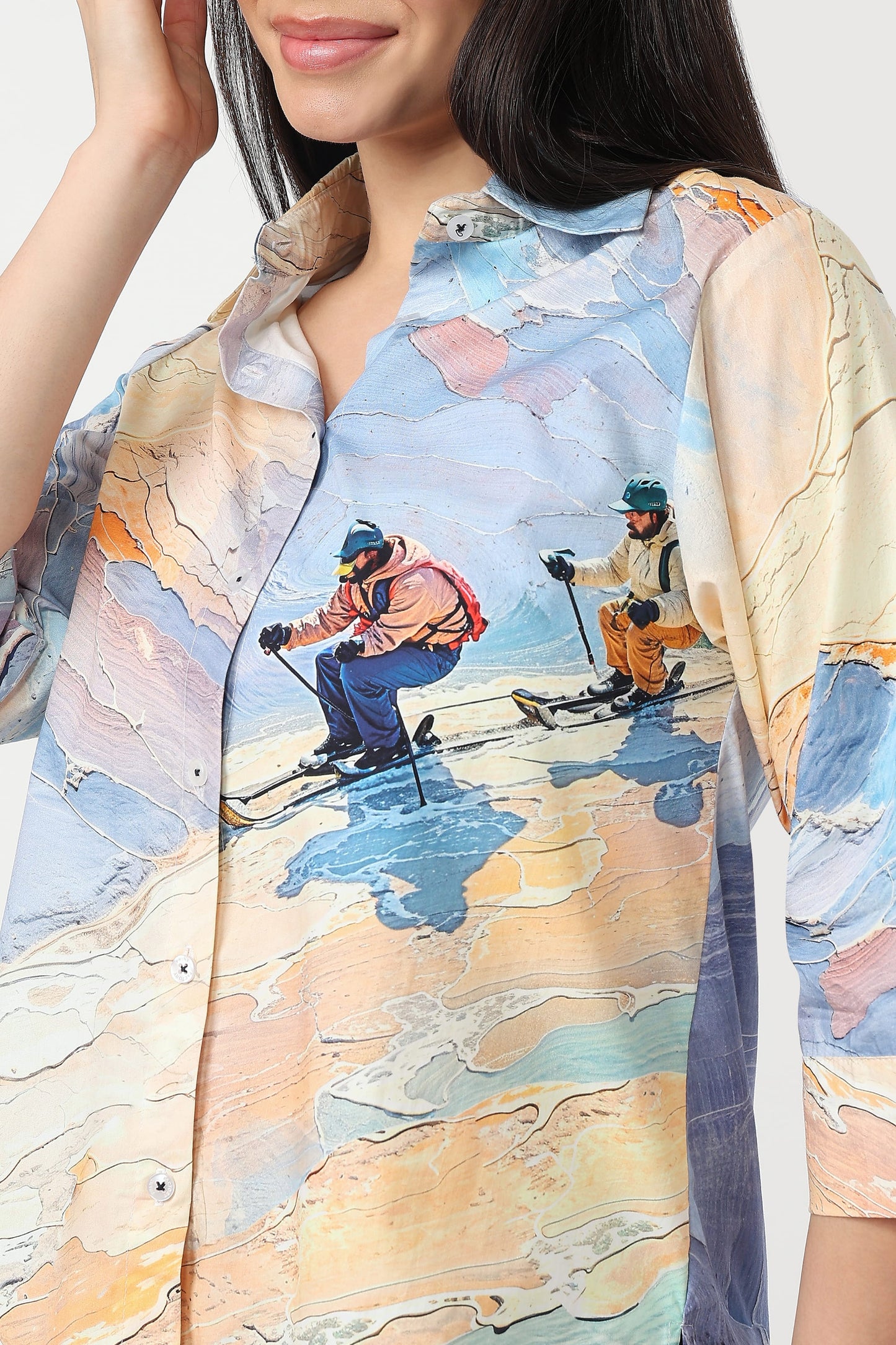 A close-up of a woman wearing a button-up shirt with a colorful painterly ski scene print. The shirt features abstract brushstroke textures in shades of blue, beige, orange, and yellow, with illustrated skiers in the center.