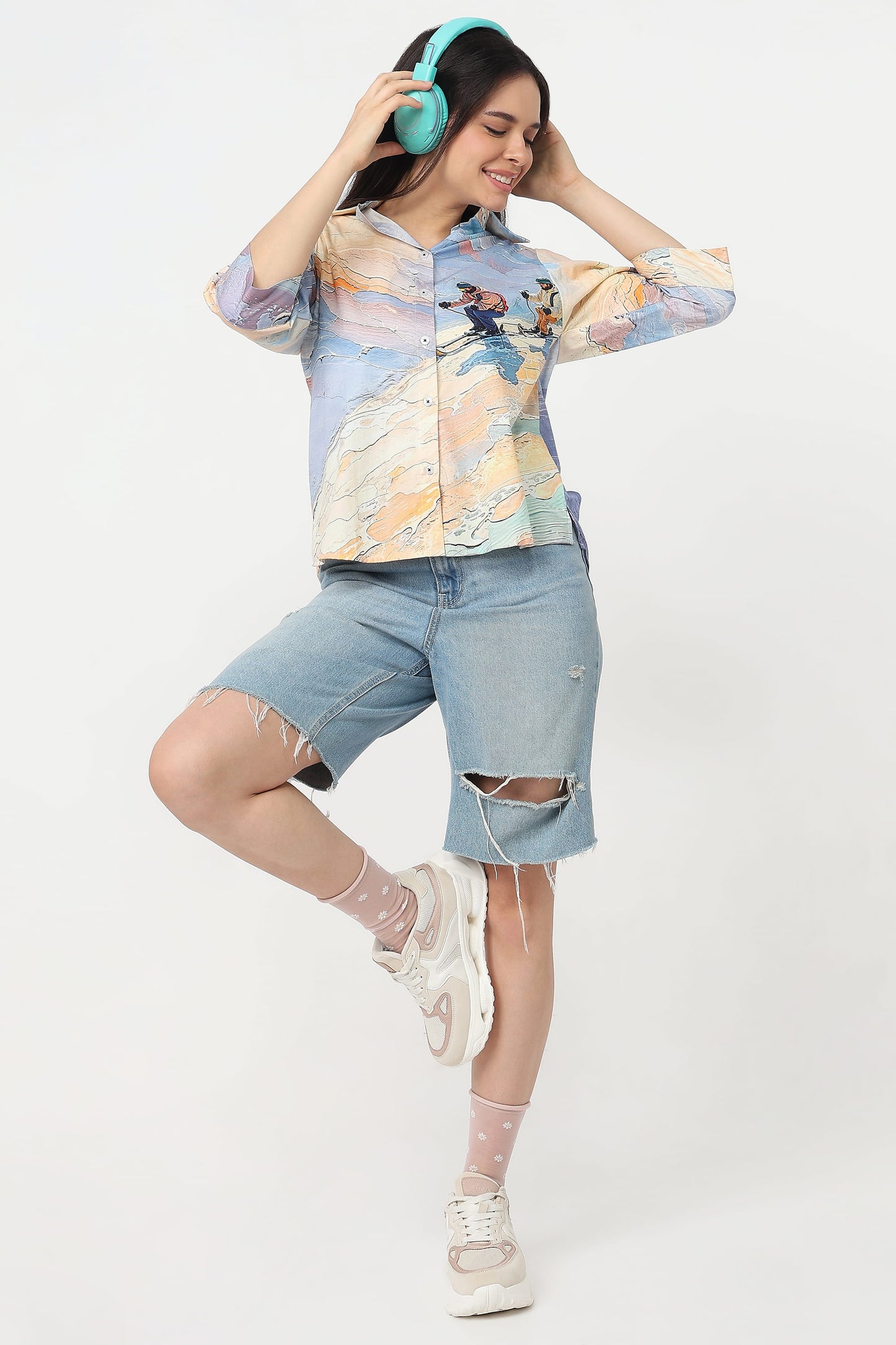 A woman posing in a studio wearing a light blue and yellow abstract print button-up shirt, distressed denim shorts, white socks, and white sneakers, while holding turquoise headphones.