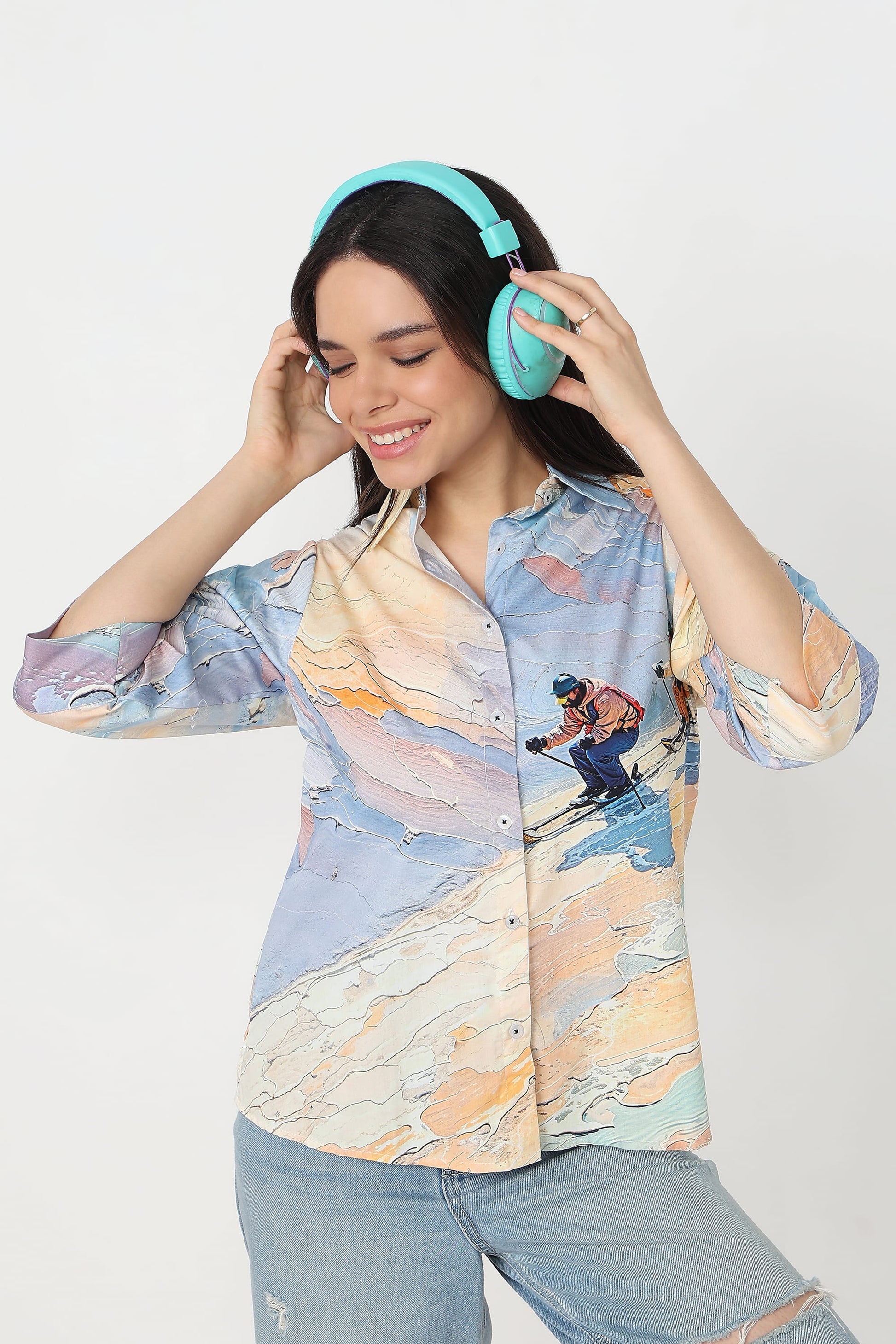 A woman wearing a light blue and beige abstract-patterned button-up shirt with a skier graphic, light-wash jeans, and bright turquoise headphones, posing against a light gray studio background.