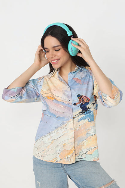 A woman wearing a light blue and beige abstract-patterned button-up shirt with a skier graphic, light-wash jeans, and bright turquoise headphones, posing against a light gray studio background.