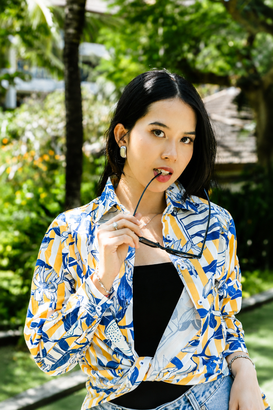 A woman wearing a vibrant blue, yellow, and white patterned button-up shirt over a black top, holding a pair of sunglasses in her hand, set against a lush green outdoor background.