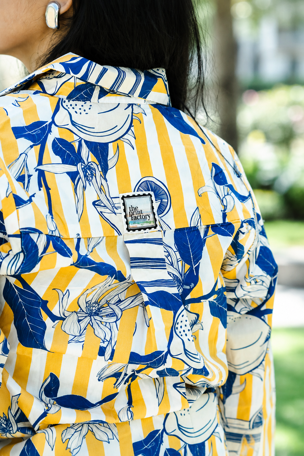 A close-up of a person wearing a yellow and white striped button-up shirt with a blue botanical print, featuring a visible 'Blue Factory' brand patch on the chest, set against a blurred outdoor background.