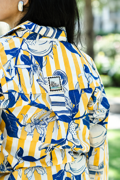A close-up of a person wearing a yellow and white striped button-up shirt with a blue botanical print, featuring a visible 'Blue Factory' brand patch on the chest, set against a blurred outdoor background.