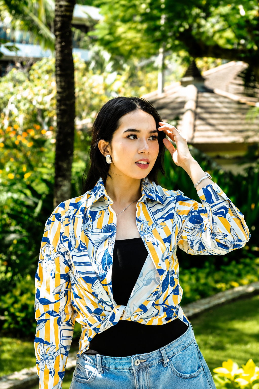 A woman wearing a blue, yellow, and white patterned button-up shirt tied at the waist over a black top and blue jeans, standing outdoors in a lush green garden.