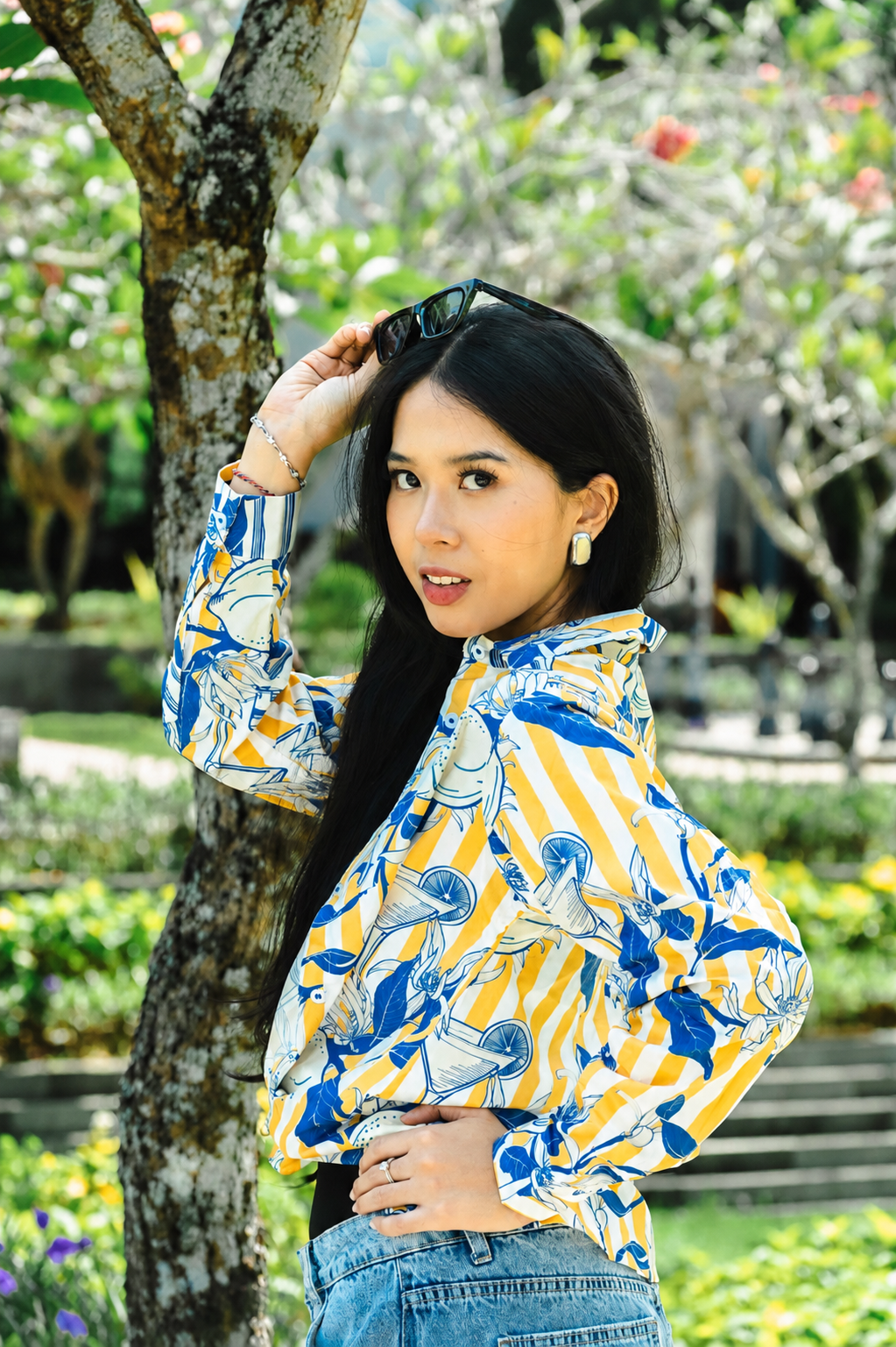 A woman posing outdoors in a vibrant yellow and blue patterned long-sleeve blouse with blue jeans, standing next to a tree in a garden setting.