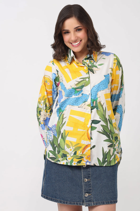 A woman modeling a long-sleeve button-up shirt with a vibrant yellow, blue, green, and white tropical graphic print, paired with a denim skirt against a plain white background.