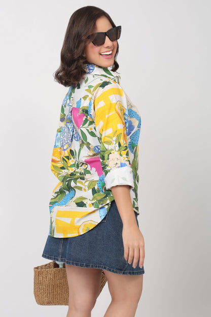 A woman wearing a colorful tropical floral print button-up shirt with yellow, blue, pink, and green patterns, paired with a denim mini skirt and sunglasses, holding a woven straw bag against a light gray background.