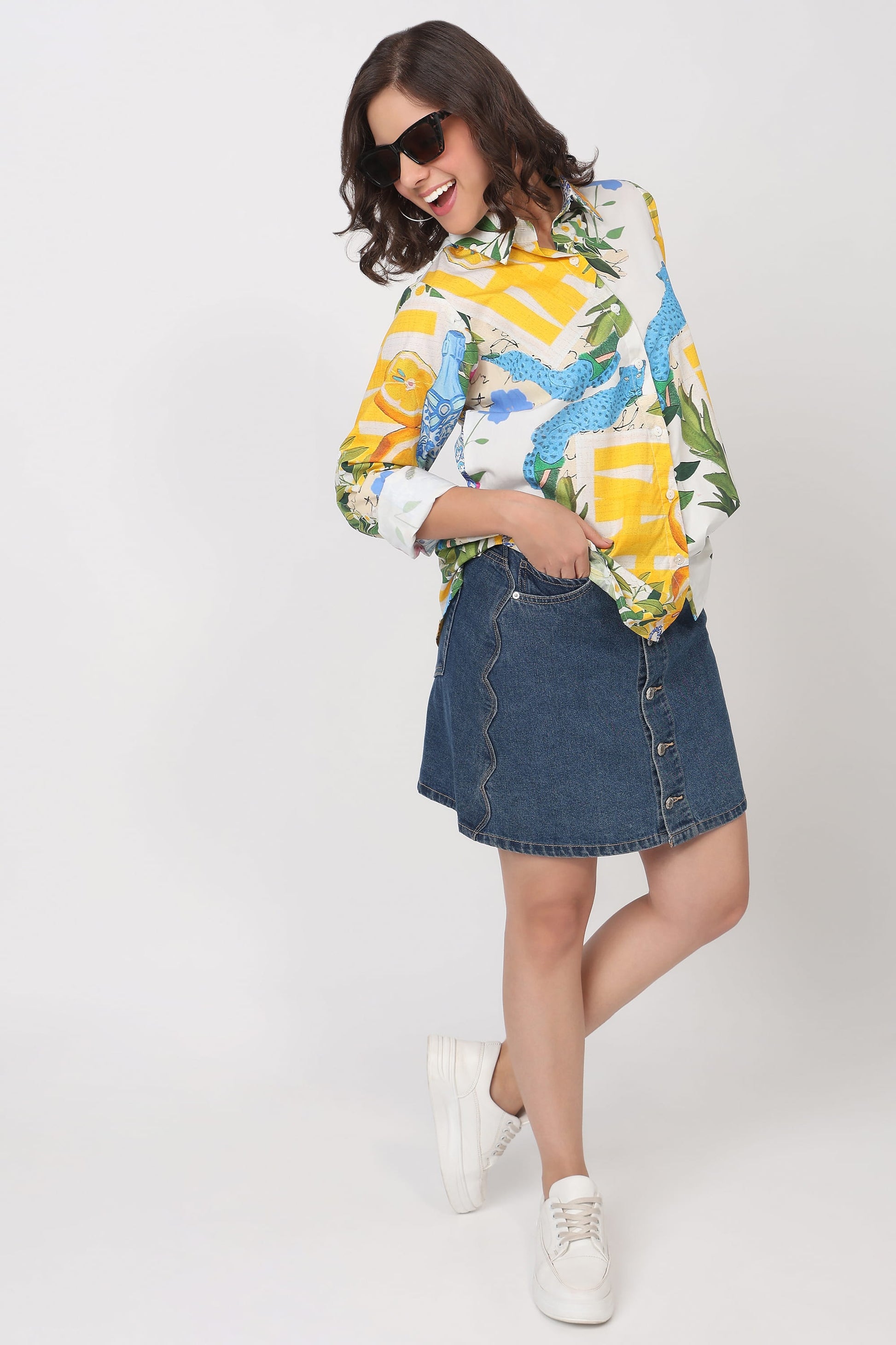 A woman modeling a colorful tropical print button-up shirt and a blue denim mini skirt, paired with white sneakers, against a plain light gray studio background.