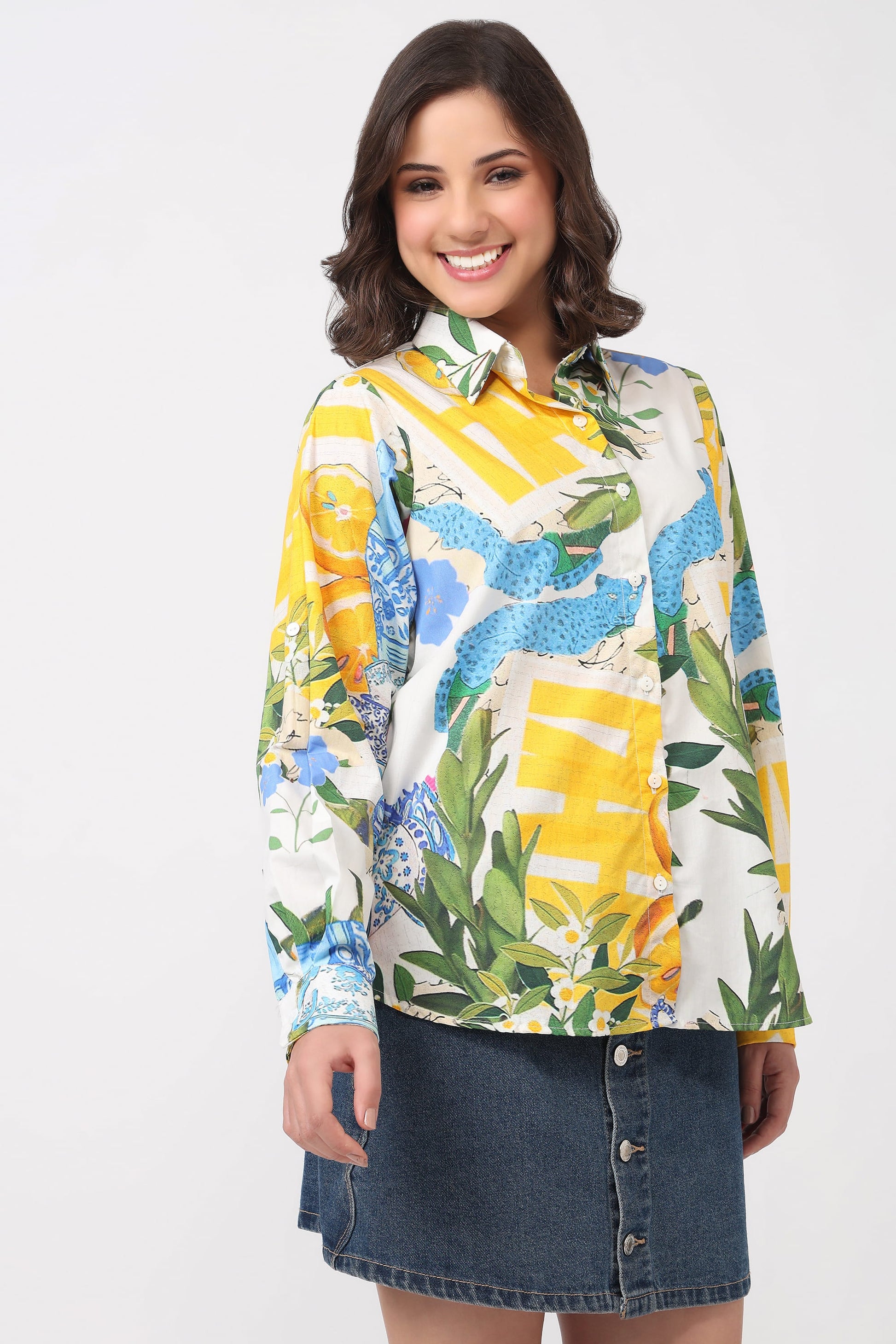 A woman modeling a long-sleeve button-up shirt with a vibrant yellow, blue, green, and white tropical floral print, paired with a dark denim mini skirt against a light gray studio background.
