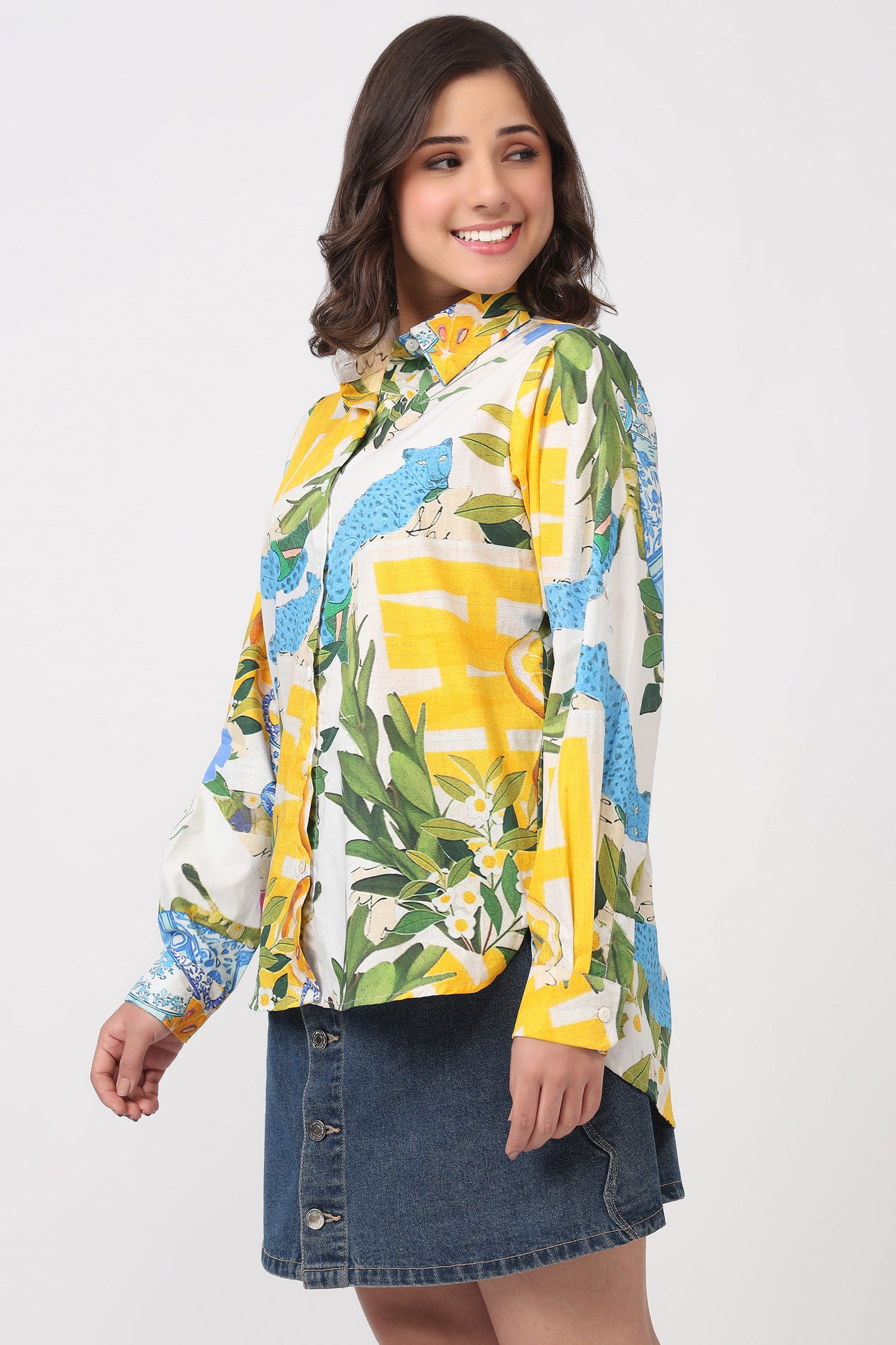 A woman modeling a long-sleeve button-up shirt with a vibrant yellow, blue, and green tropical and abstract print, paired with a denim skirt against a plain white studio background.
