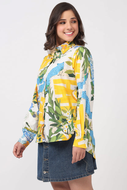 A woman modeling a long-sleeve button-up shirt with a vibrant yellow, blue, and green tropical and abstract print, paired with a denim skirt against a plain white studio background.