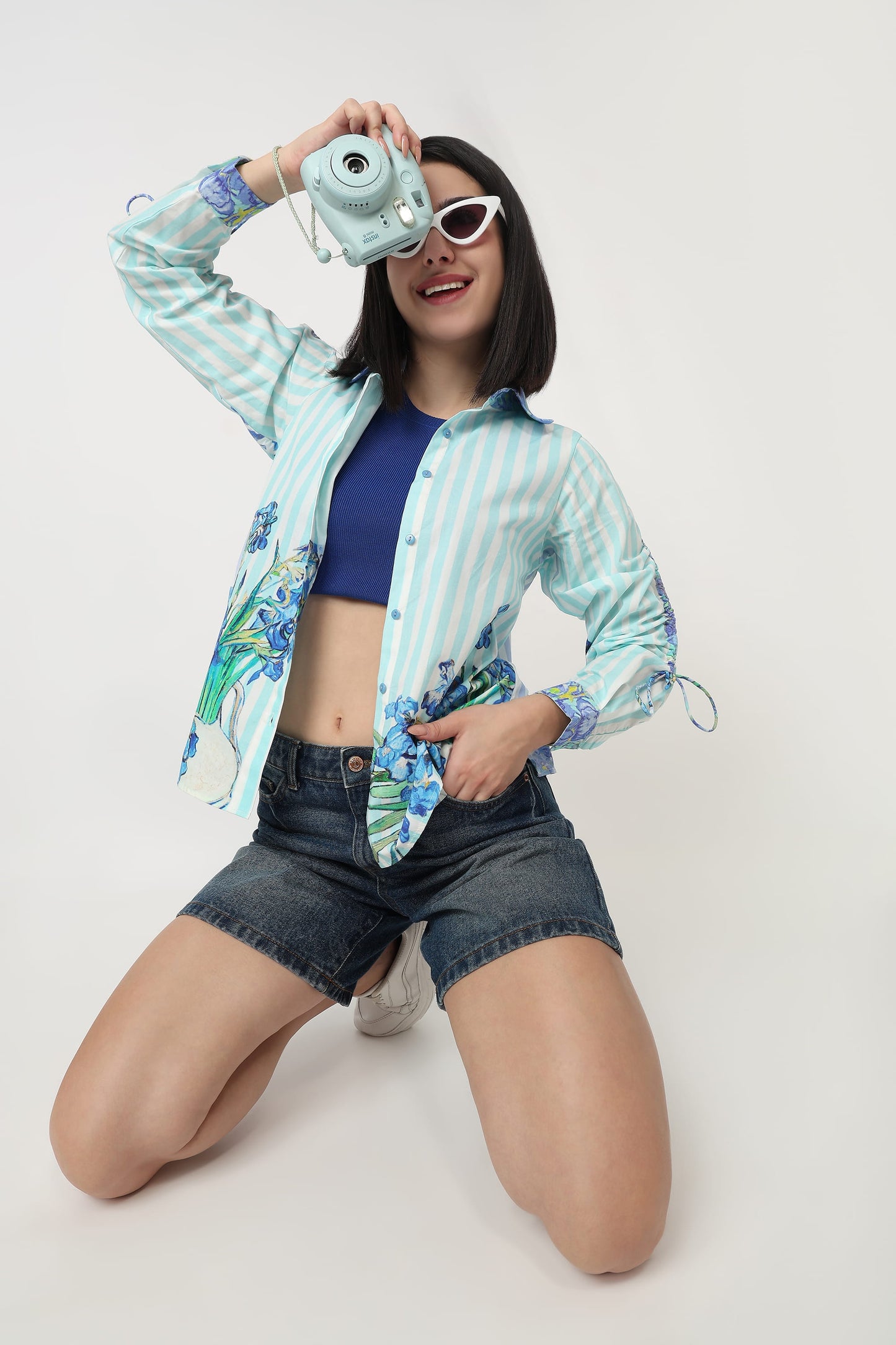 A woman kneels on a light gray studio floor wearing a light blue and white striped button-up shirt with a blue floral print on the sleeves and hem, a navy blue crop top, denim shorts, white sunglasses, and holding a mint green instant camera to her eye.