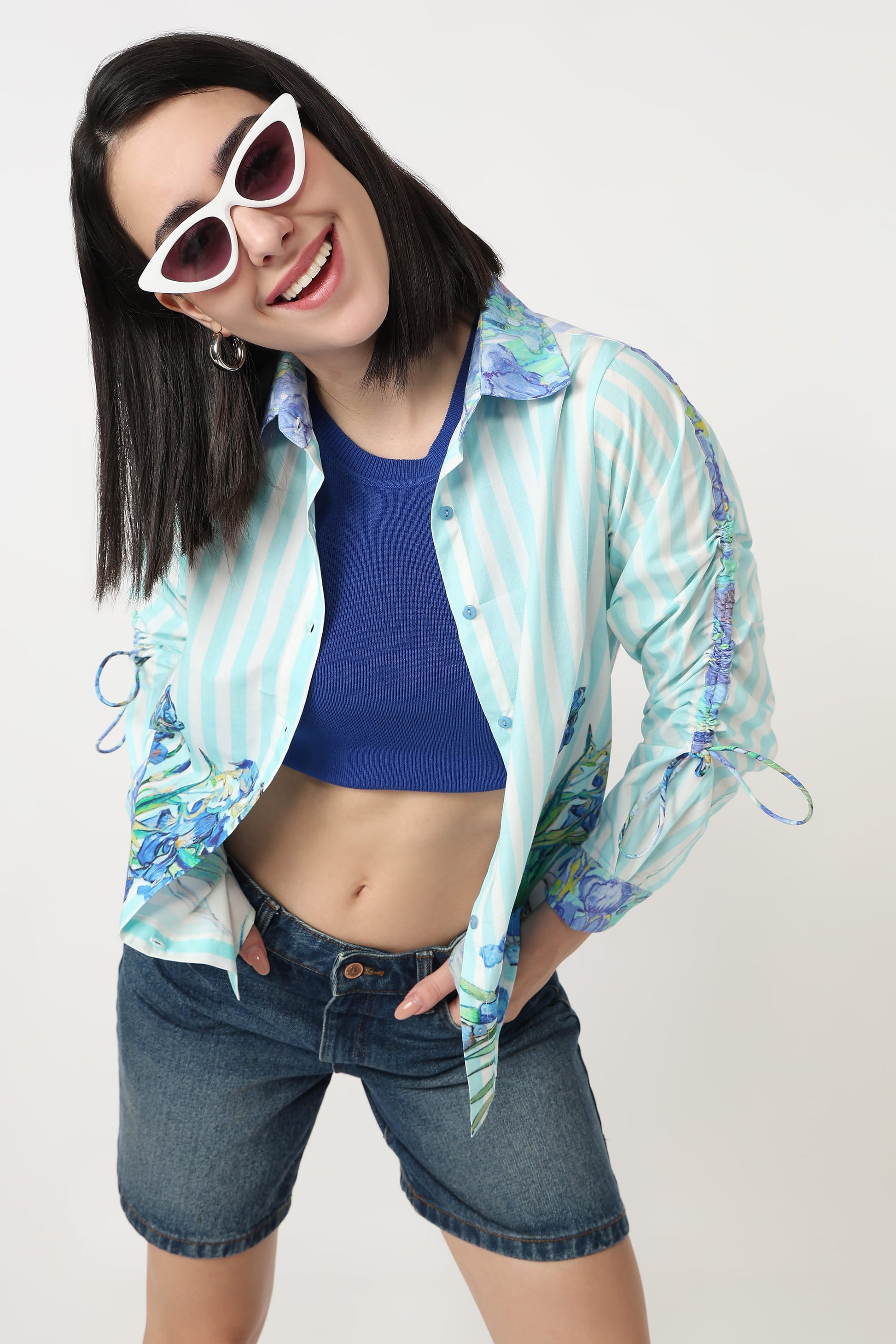 A woman modeling a light blue and white vertically striped button-up shirt with blue and green floral patterns on the sleeves and cuffs, worn open over a blue crop top and denim shorts.