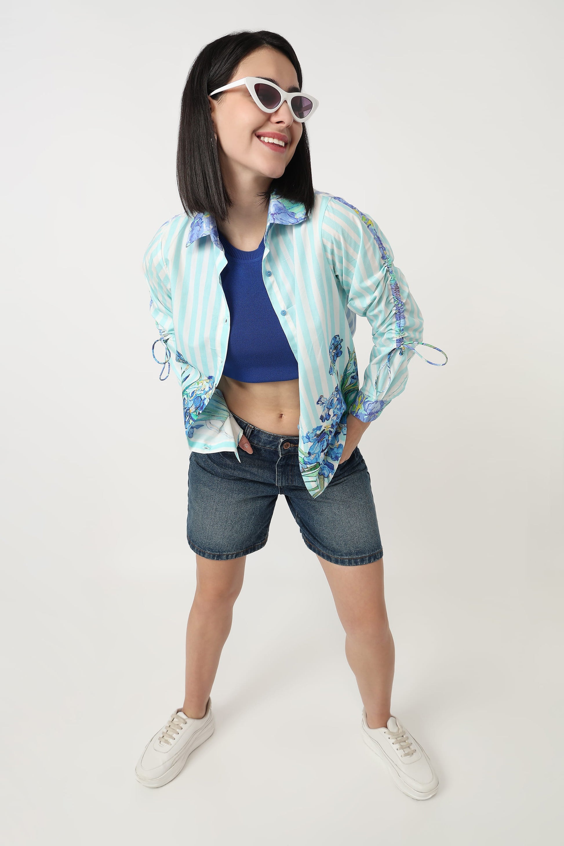 A woman modeling a light blue and white striped bomber jacket with blue floral patterns on the sleeves, worn over a blue crop top and dark denim shorts, on a white studio background.