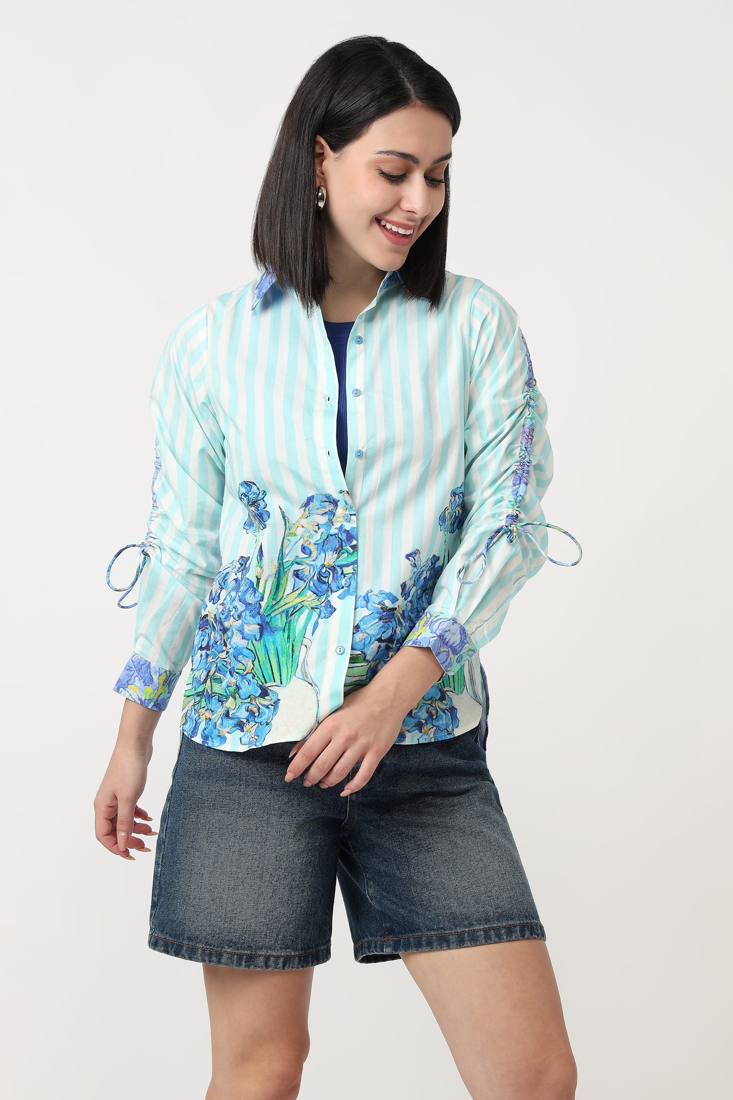 A woman modeling a light blue and white striped button-up shirt with blue floral prints on the front and sleeves, paired with dark denim shorts, against a plain light background.