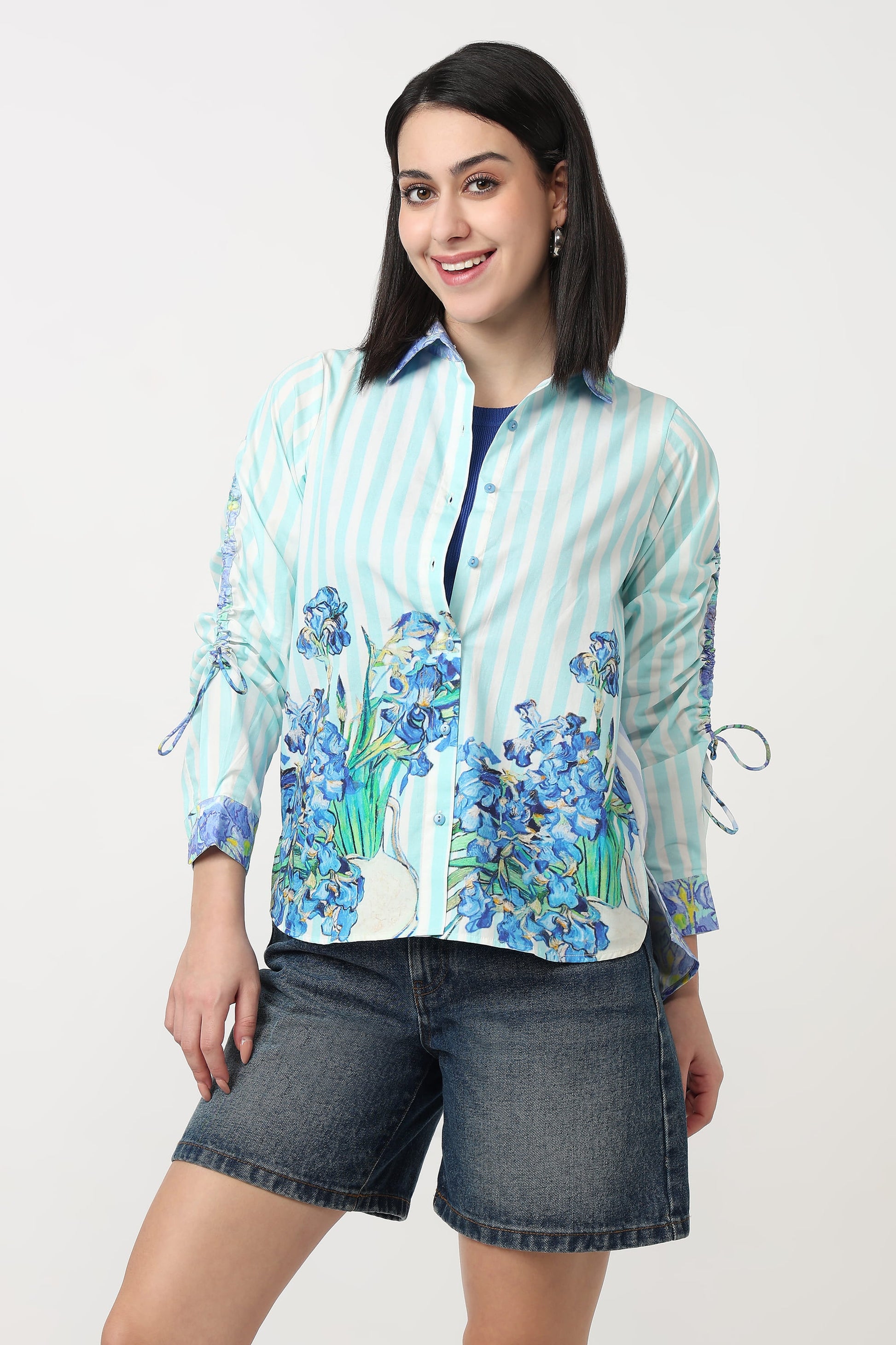 A woman modeling a light blue and white striped button-up blouse with blue floral prints on the front and sleeves, paired with dark denim shorts, against a plain light gray background.