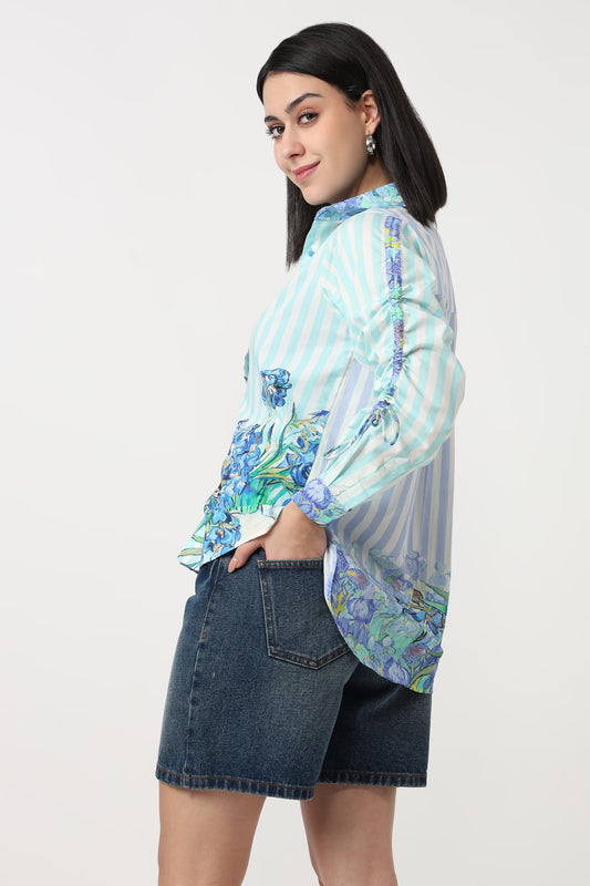 A woman modeling a light blue and white striped long-sleeve blouse with blue and green floral print details on the sleeves and hem, paired with denim shorts against a plain white background.