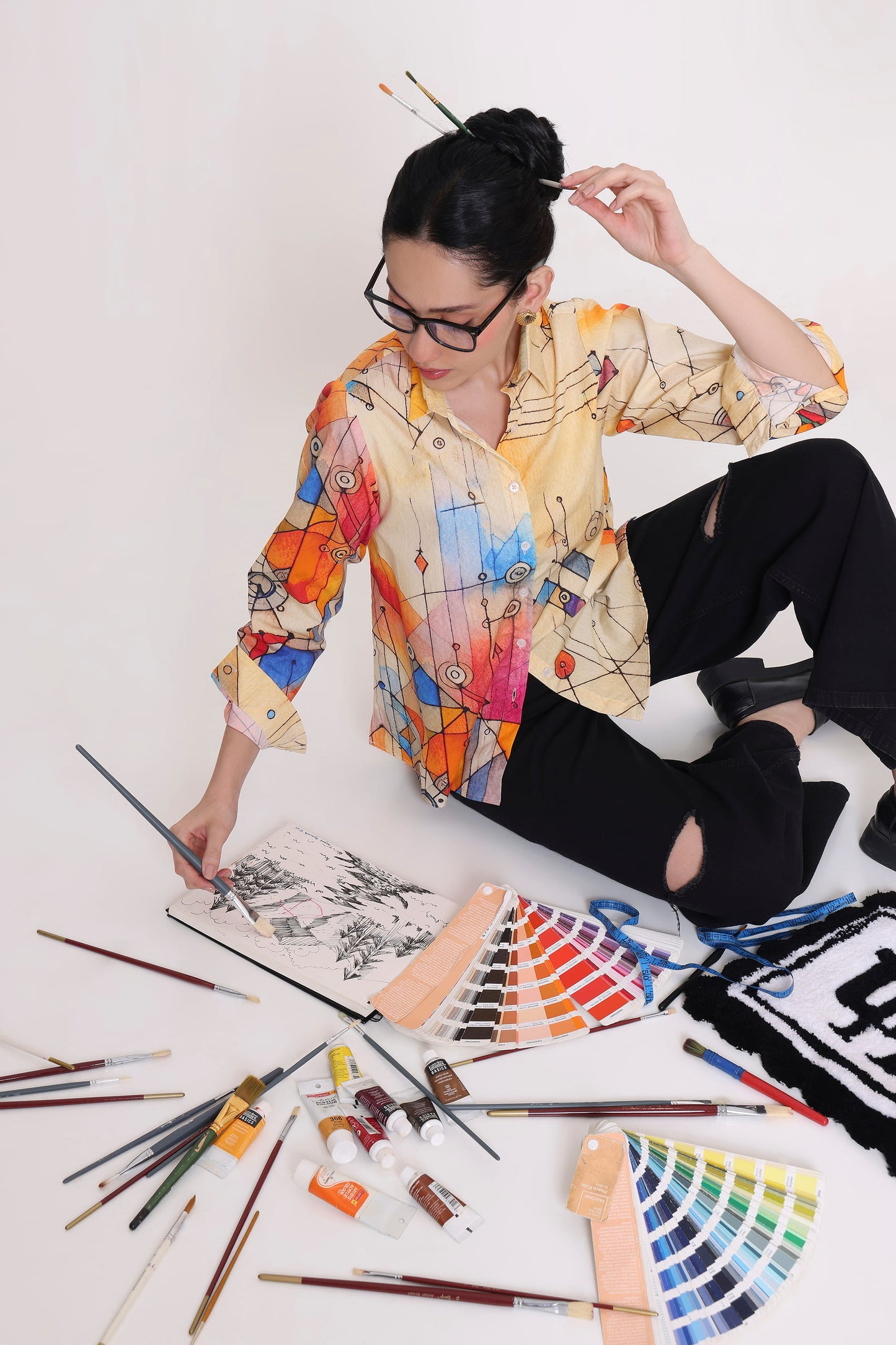 A woman with glasses and her hair in a bun sits on a white floor surrounded by painting supplies, including paintbrushes, paint tubes, a palette of color swatches, and a canvas. She is wearing a colorful abstract print button-up shirt and black ripped jeans.