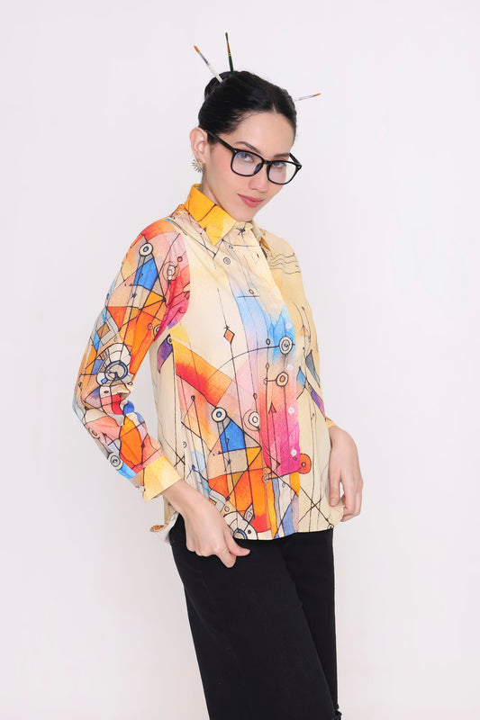 A woman wearing a long-sleeve button-up shirt with a vibrant multicolor abstract geometric print on a cream base, paired with black pants and glasses, posing against a plain white background.