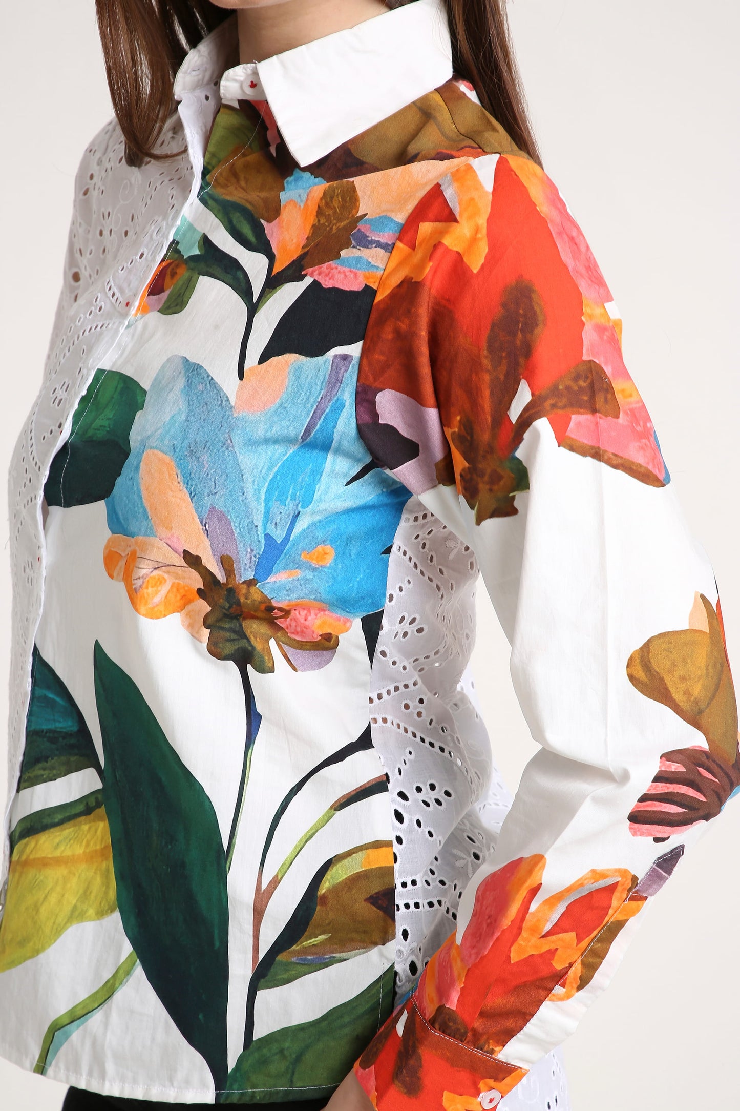A close-up of a woman wearing a white long-sleeve blouse with a vibrant, painterly floral print in orange, blue, green, and brown, featuring eyelet lace details.