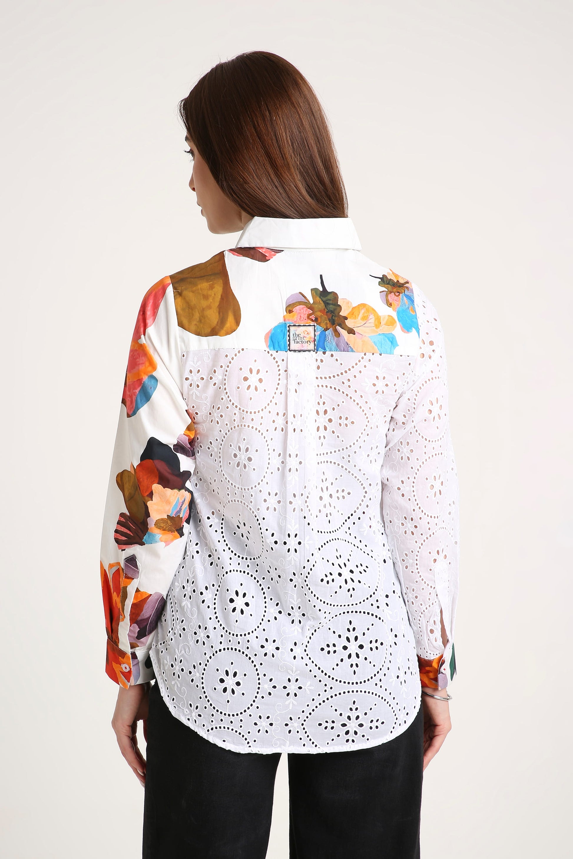 A woman shown from the back wearing a white long-sleeve button-up shirt with colorful abstract painterly prints on the shoulders and sleeves, paired with black pants, against a plain light background.