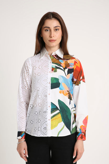 A woman wearing a long-sleeve button-up shirt with a split design: one half is white eyelet lace, and the other half features a colourful tropical floral print on a white base, paired with black pants against a light grey studio background.