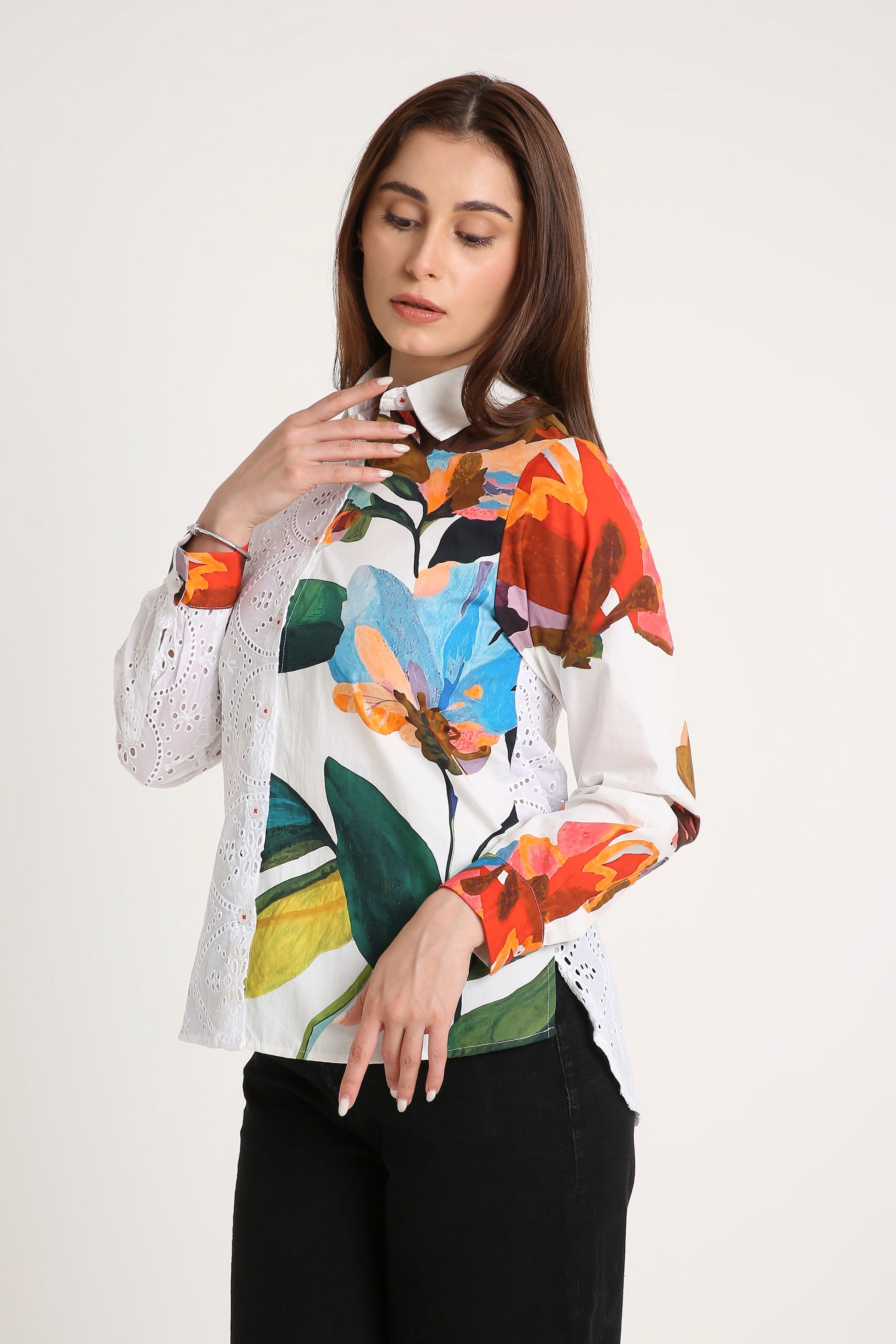 A woman wearing a white long-sleeve blouse with a vibrant multicolor floral print and sheer white lace sleeves, paired with black pants, against a plain light background.