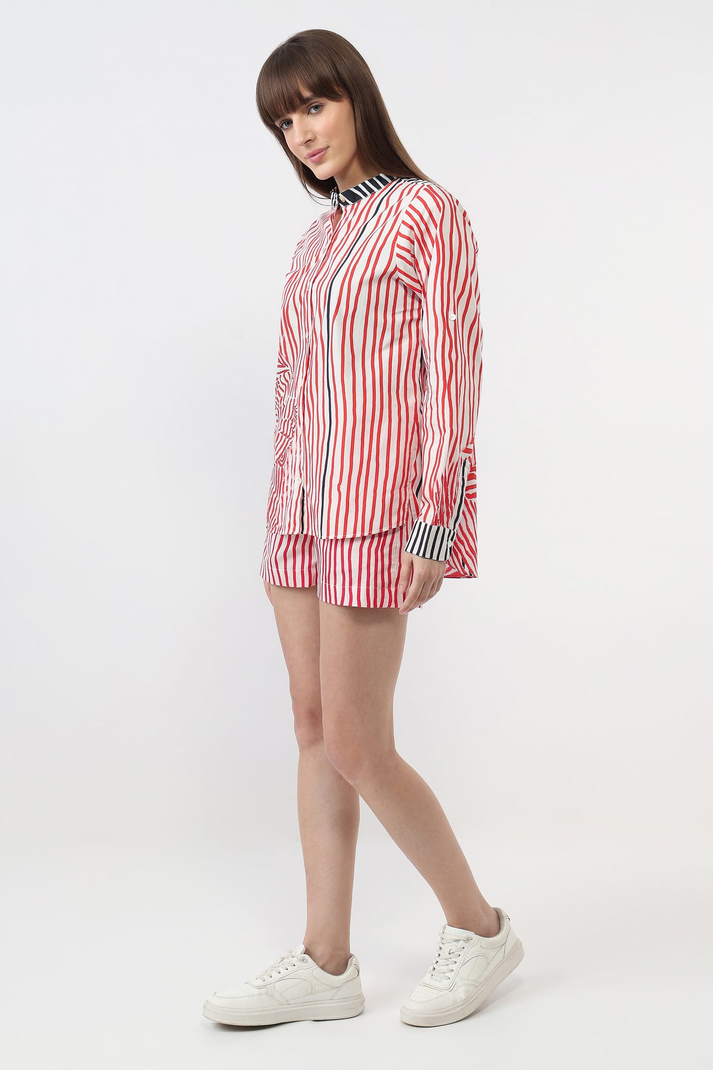 A woman modeling a matching red, white, and blue striped button-up shirt and shorts set, paired with white sneakers, against a plain white studio background.